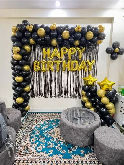 A classic birthday decoration for a living room, featuring a black and gold balloon arch over a black fringe curtain.