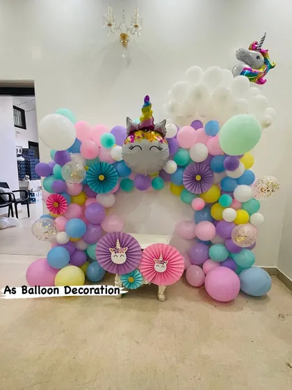 A magical unicorn-themed party backdrop, perfect for a child's birthday or a baby welcome. This setup includes a multi-colored pastel balloon arch, a large unicorn foil balloon, and charming paper fan details.