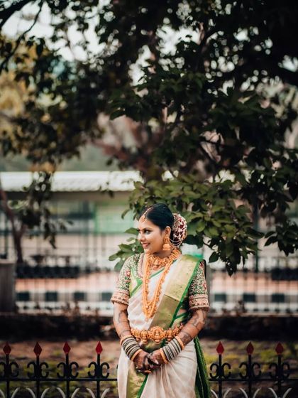 A classic portrait of a South Indian bride in a beautiful white and green saree, standing gracefully in a garden setting.