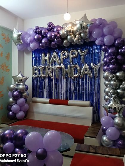 A purple and silver balloon arch with a blue fringe curtain and "Happy Birthday" foil letters.