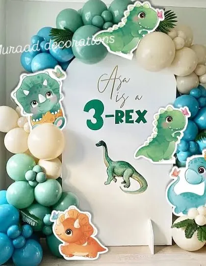 A playful '3-Rex' dinosaur theme for a third birthday. The decoration features cute cartoon dinosaur cutouts on a simple white backdrop, framed by a balloon garland in shades of teal, blue, and cream.