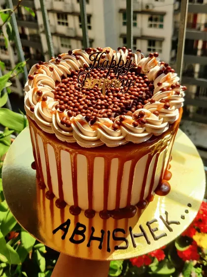 Living the sweet caramel life. This tall cake features a caramel drip, swirls of caramel buttercream, and a topping of crunchy chocolate balls, a delight for anyone who loves caramel.