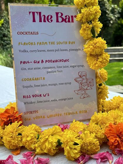 A custom bar menu for the Mehendi, featuring creative cocktails like the 'Coorgarita', framed with fresh marigold flowers.