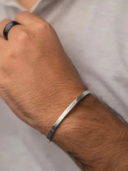 A man's wrist with a silver cuff engraved with 'AVIRAJ'. The bracelet's sleek design is perfect for stacking or wearing alone as a statement piece.