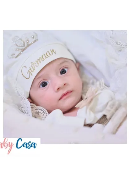 A beautiful close up of a newborn wearing a personalized cap for "Gurmaan." The gold embroidery on the white cap adds a touch of personalized luxury.