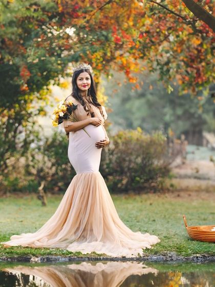 An expectant mother in a lovely mermaid-style gown stands by a pond in a park with autumn foliage. The warm colors of the background beautifully complement her gentle and radiant look.