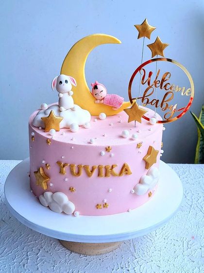 A "Welcome Baby" cake with a celestial theme. This pink cake features a fondant moon, stars, clouds, and a cute sleeping baby and bunny topper.