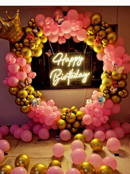 A glamorous pink and gold birthday decoration. This full balloon ring is adorned with a crown, butterfly accents, and a 'Happy Birthday' neon sign, with balloons covering the floor.