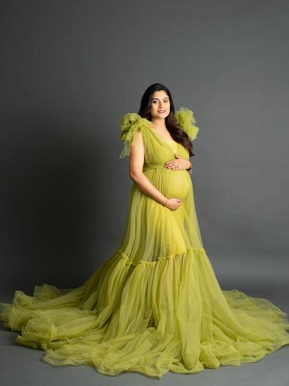 A graceful portrait of an expecting mother in a chartreuse green tulle gown. She stands against a simple grey background, her hands cradling her bump, showcasing the dress's ruffled shoulders.