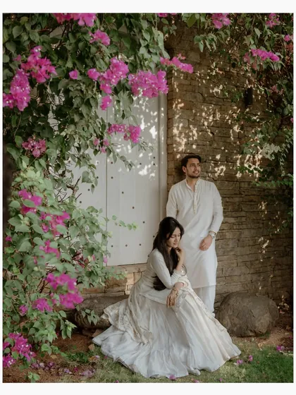 A stylish portrait of a couple against a stone wall adorned with vibrant pink bougainvillea. Their coordinated white outfits and poised expressions create a chic and modern pre-wedding photo.