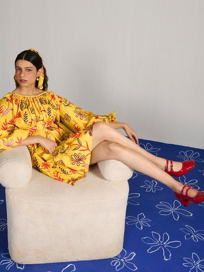 A relaxed, seated pose from the Joskai Studio shoot. The styling of the yellow printed dress with bold red shoes creates a fun and confident color-blocked look.