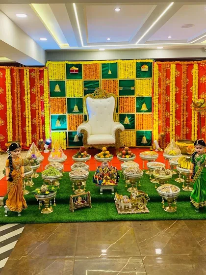 A full view of a traditional Seemantha setup with a green lawn, offering trays, and a colorful checkerboard backdrop. The hanging garlands and traditional dolls complete this festive scene.