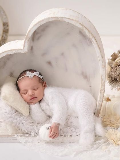 A newborn in a fluffy white outfit sleeps inside a large, white heart-shaped bowl, surrounded by white pumpkins and decor for an all-white theme.