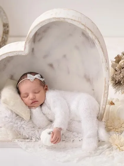 A newborn in a fluffy white outfit sleeps inside a large, white heart-shaped bowl, surrounded by white pumpkins and decor for an all-white theme.