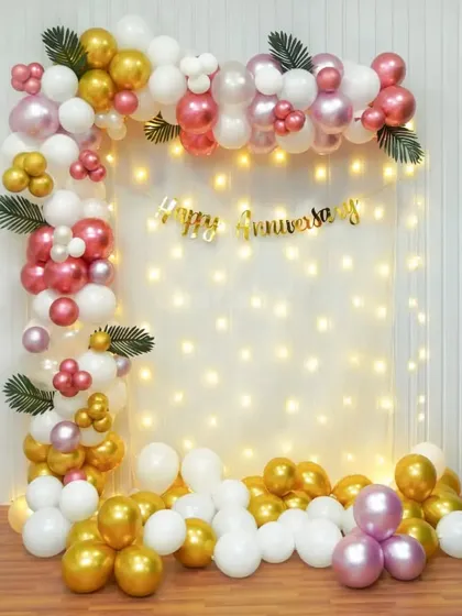A simple and elegant anniversary photo corner with a balloon garland in pink, gold, and white against a fairy light wall.