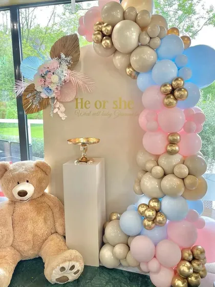 A sophisticated gender reveal setup using a neutral backdrop and a beautiful balloon arch with pastel pink, blue, and champagne gold colors. The large teddy bear adds a soft and cuddly element to the design.