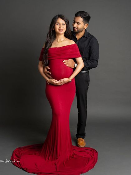 A romantic pose showing a couple's connection. The partner embraces the mom-to-be from behind, both cradling her belly, in a classic red gown and black shirt combination.