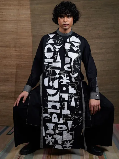 A full-length shot of the model seated, showcasing the entire abstract-faced tunic. The composition is strong and balanced, presenting the garment as a piece of wearable art.