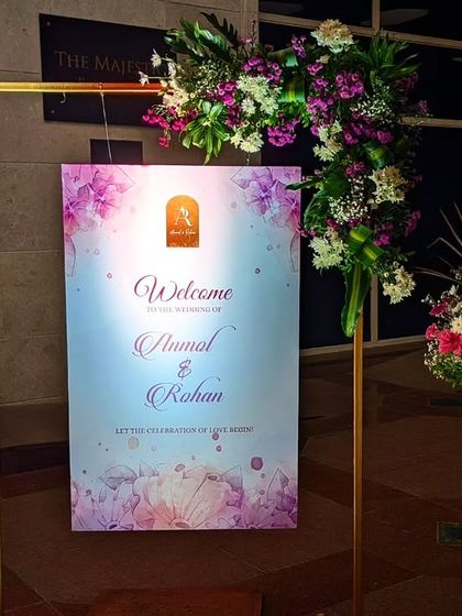 A simple yet elegant welcome sign for Anmol and Rohan, framed with a modern golden structure and a beautiful arrangement of purple and white flowers.