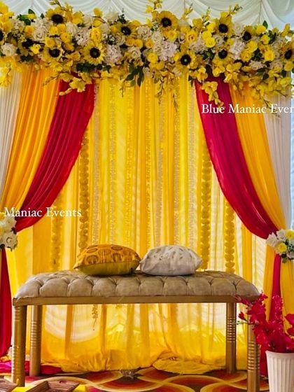 A bright and sunny Haldi/Mehendi backdrop with yellow, pink, and red drapes and a sunflower floral arrangement.