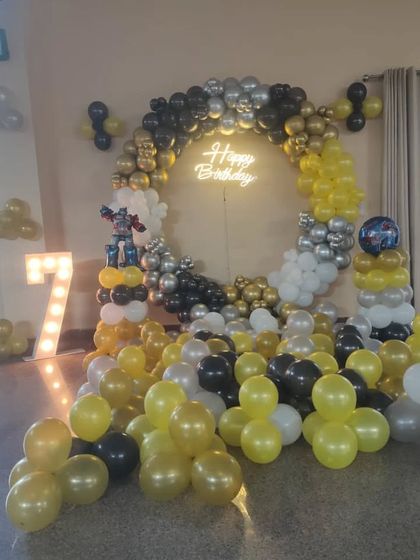 A fun 7th birthday setup with a Transformers theme, using a yellow, black, and silver balloon ring, a neon sign, and a light-up number '7'.