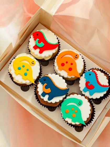 A box of six dinosaur-themed cupcakes, perfect for a kid's birthday party. Each cupcake features a brightly coloured, 2D fondant dinosaur topper in red, yellow, orange, blue, and green, resting on a bed of white buttercream.