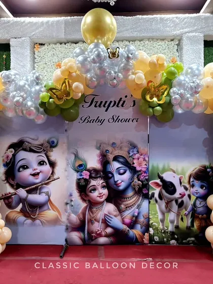 A baby shower stage featuring a Radha Krishna theme with a custom backdrop. The balloon arch uses a unique color palette of green, peach, and gold.