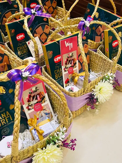 A side view of a row of identical wedding favor baskets. Each is consistently decorated with purple ribbons and flowers and filled with a box of Chana Burfi, highlighting the uniformity and elegance of my bulk preparations.