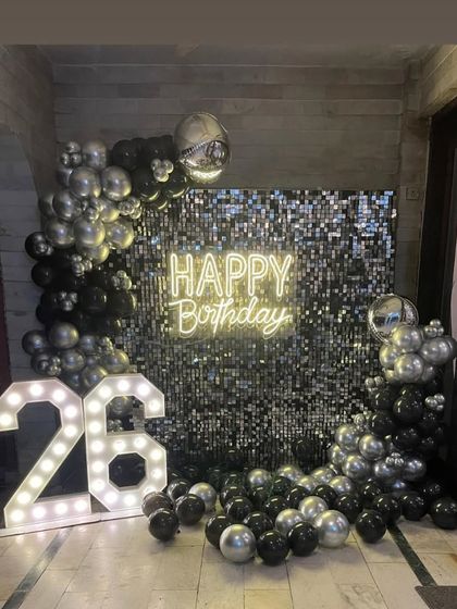 A glamorous 26th birthday setup featuring a silver sequin backdrop, a "Happy Birthday" neon sign, and a black and silver balloon garland.