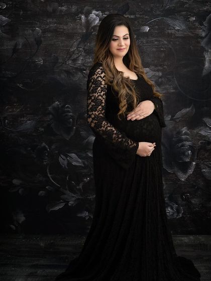A classic maternity portrait in a black lace gown. The dark floral backdrop adds a touch of gothic romance and texture, resulting in a timeless and elegant image.