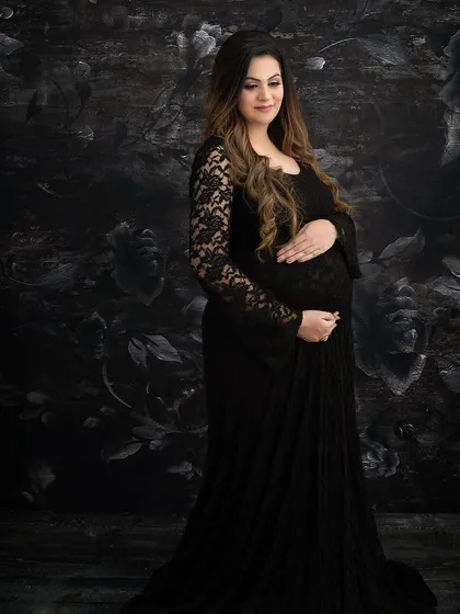 A classic maternity portrait in a black lace gown. The dark floral backdrop adds a touch of gothic romance and texture, resulting in a timeless and elegant image.