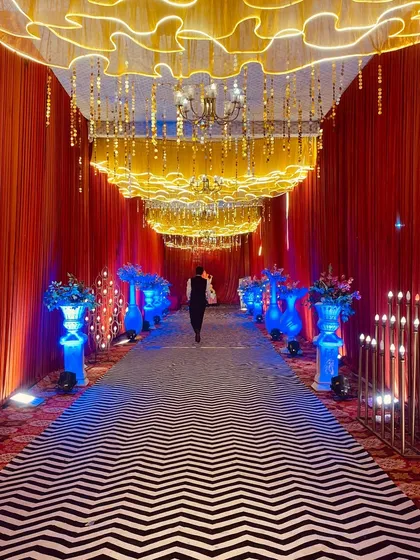 A modern and chic entrance with a black and white chevron carpet. The walkway is illuminated by blue uplighting on the vases and a unique wavy, lit ceiling treatment.