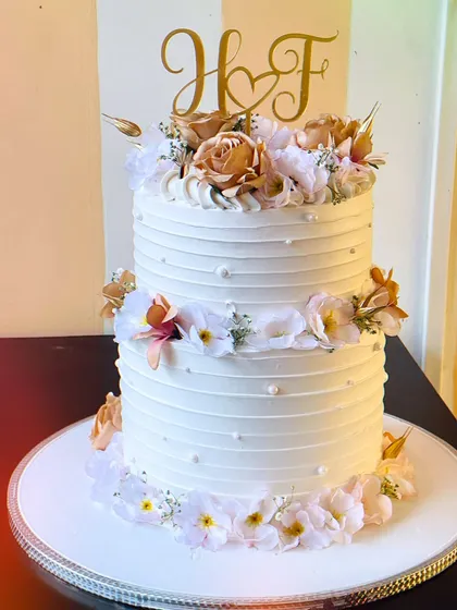 A different perspective of the elegant two-tier wedding cake, showing the beautiful initial topper and the careful placement of each flower. This cake is made with premium cream cheese frosting and white chocolate.