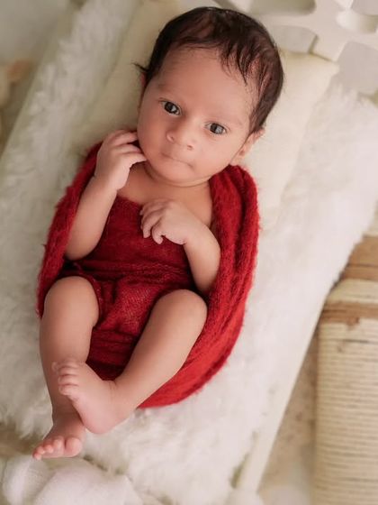 An awake newborn wrapped in red looks directly at the camera while lying on a tiny white bed with prop blocks showing the date.