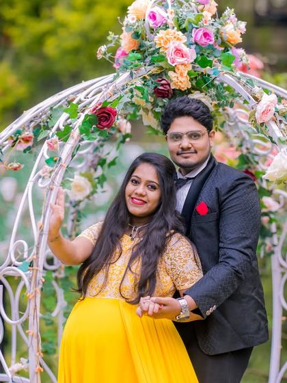A happy maternity portrait in front of a white floral archway, with the garden's pink and red flowers creating a soft and beautiful background.