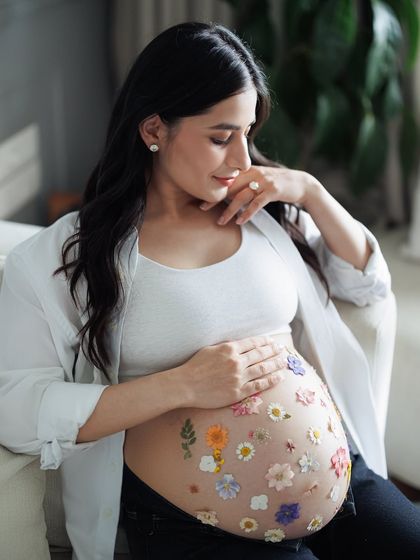 A quiet moment of reflection for the mother-to-be. Seated comfortably on a couch, she looks down at her flower-adorned belly with a gentle smile.