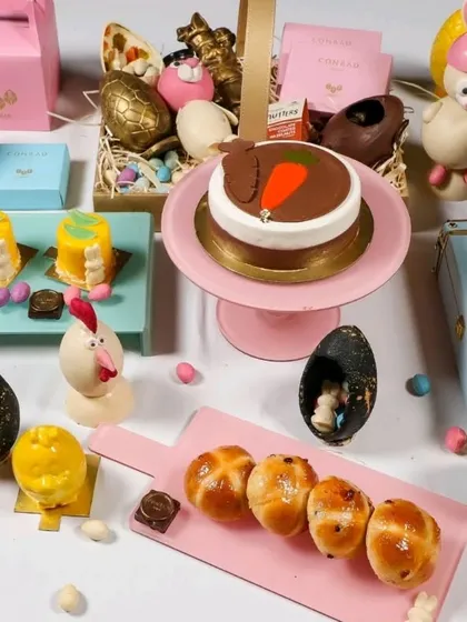 A beautiful Easter-themed high tea spread, featuring cakes, pastries, hot cross buns, and chocolate eggs, all presented on colorful stands.