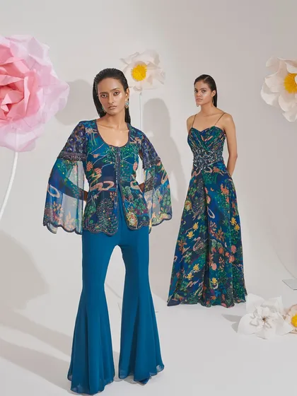A dual-model shot showcasing two different ways to wear a contemporary blue print. One model wears a jumpsuit, while the other is in a flared pants set with a bell-sleeve jacket, demonstrating the range of a cohesive collection.