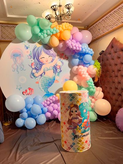 A cute mermaid theme decoration using a round backdrop with a mermaid illustration and a pastel rainbow balloon garland.