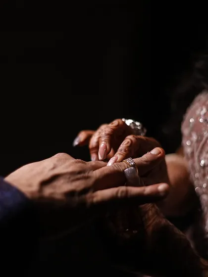 A close-up, detailed shot of the ring exchange, focusing on the hands and the ring as a symbol of commitment. The dramatic lighting adds a cinematic touch.