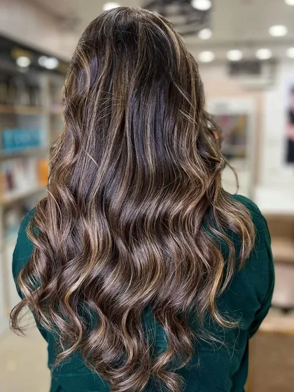 A beautiful example of balayage on long, wavy hair, seen from the back. The salon's blurred background keeps the focus on the stunning hair.