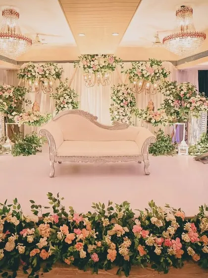 We design elegant and classic stage decor for receptions and engagements. This setup features a beautiful white chaise lounge against a backdrop of lush white and pastel pink floral arrangements, creating a sophisticated and romantic atmosphere.