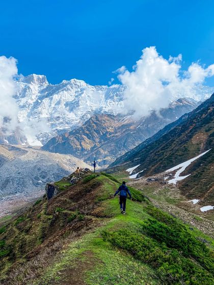 A trekker walking along a vibrant green ridge with the majestic, snow-covered Himalayan range in the background. This image captures the feeling of freedom and scale that mountain adventures provide.