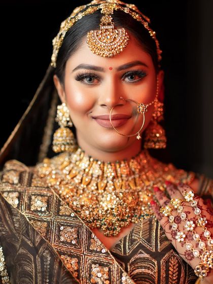 A smiling portrait of a bride in a stunning black and gold lehenga. Her makeup features a smokey eye, a warm, glowing complexion, and a nude lip, complemented by heavy gold jewelry.