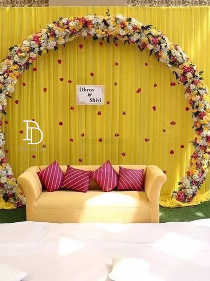 A simple yet elegant mehndi backdrop featuring a large floral ring against a bright yellow curtain. The sofa provides comfortable seating, and the setup can be personalized with the couple's names.