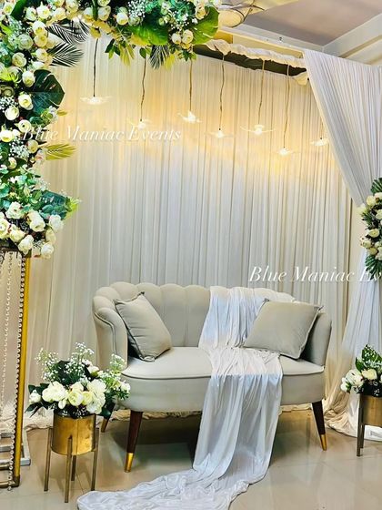 A sophisticated Roka setup with a C-shaped arch of white flowers and tropical leaves. The hanging lights and gold-accented floral stands add a touch of luxury and modernity.