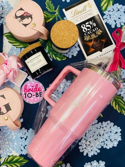 A flat lay of a gorgeous bridesmaid hamper. It includes a personalised pink glam tumbler, a floral initial jewellery box, a 'Bride to-Be' badge, a scented candle, and gourmet chocolate.