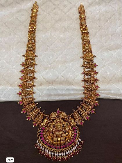This long temple haram is made from 92.5 silver with gold plating and features a repeating goddess motif. The necklace is adorned with ruby-colored stones for a rich, royal look.