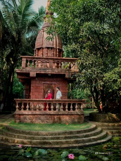 A stunning wide shot of the couple standing on the balcony of a beautiful red stone temple, surrounded by lush trees and a lily pond. A truly epic and cultural pre-wedding photo.