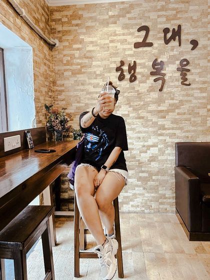 A fun, 'no-face' shot at a Korean cafe in Kolkata, holding up a drink. The Korean text on the wall adds to the authentic atmosphere.